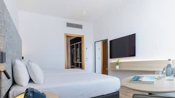 Una habitación de hotel moderna con cama, mesita de noche, silla, televisor y una pequeña mesa con un libro y vasos.
