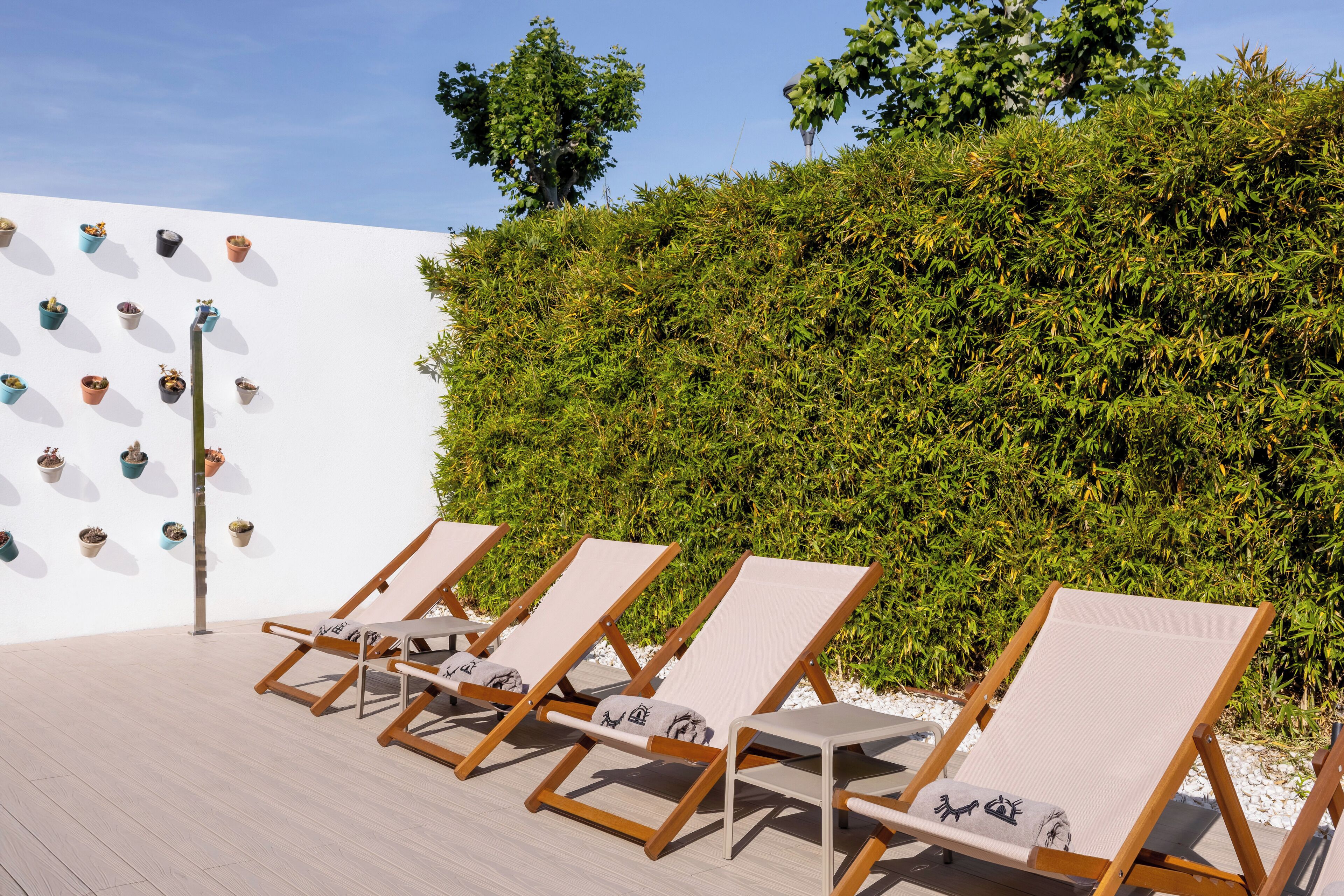 A row of wooden lounge chairs with cushions, aligned against a white wall with potted plants.