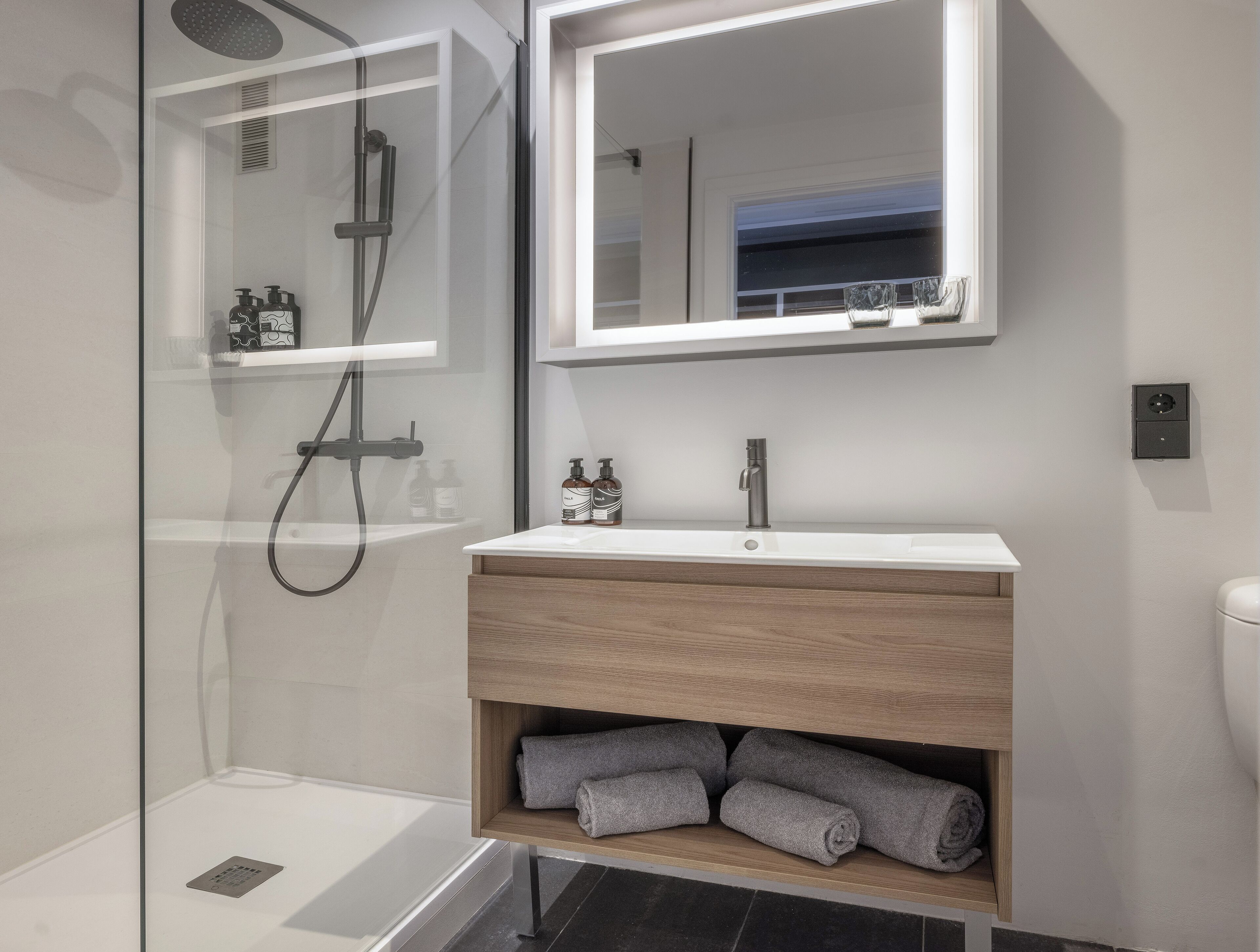 Un baño moderno con ducha envuelta en vidrio, un lavabo blanco con tocador de madera y un espejo sobre el tocador.