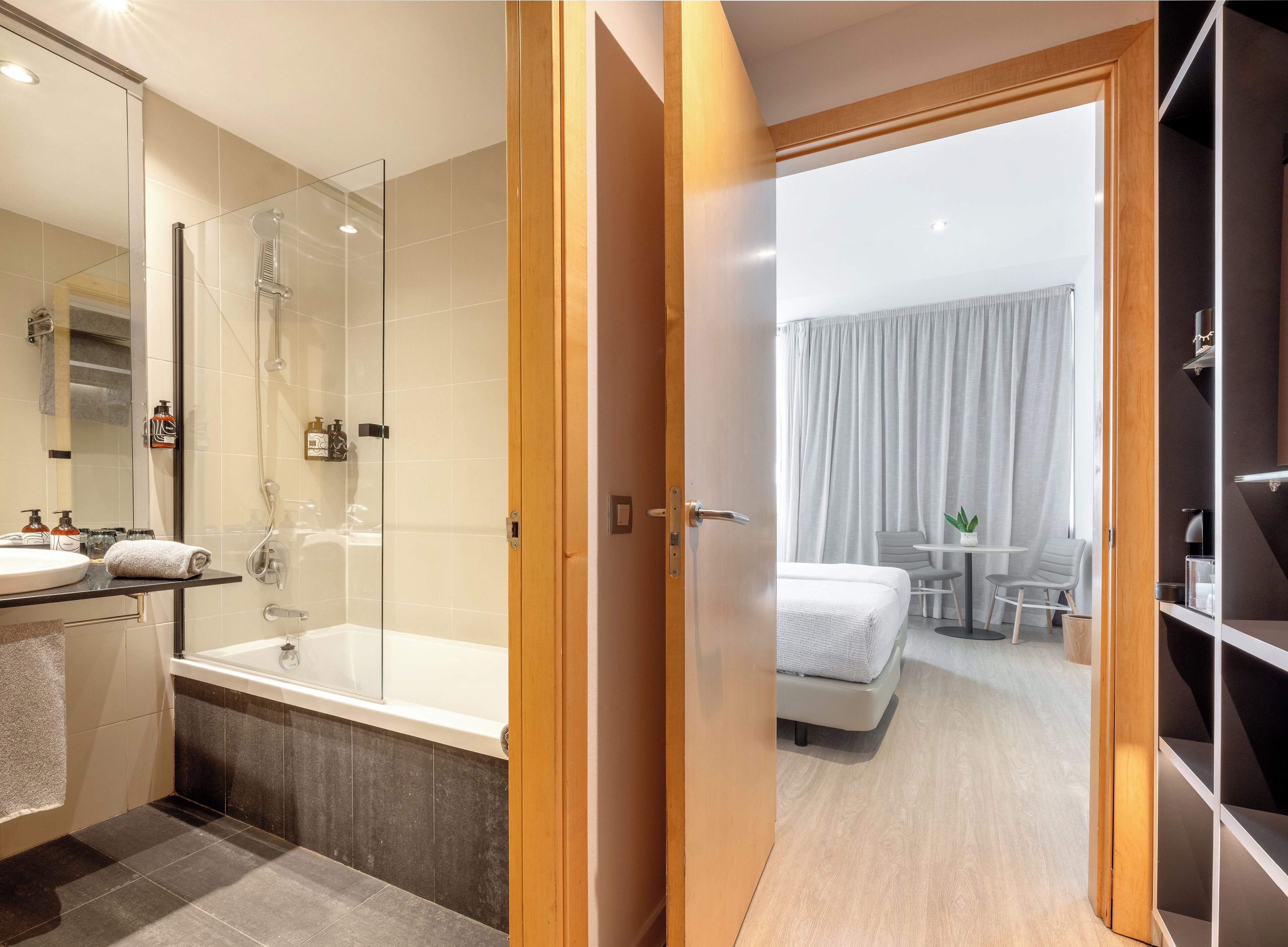 Une chambre d’hôtel moderne dotée d’une douche en verre, d’une salle de bain séparée avec une baignoire, d’un lit avec une couette blanche, d’une petite table avec une plante et d’une armoire.
