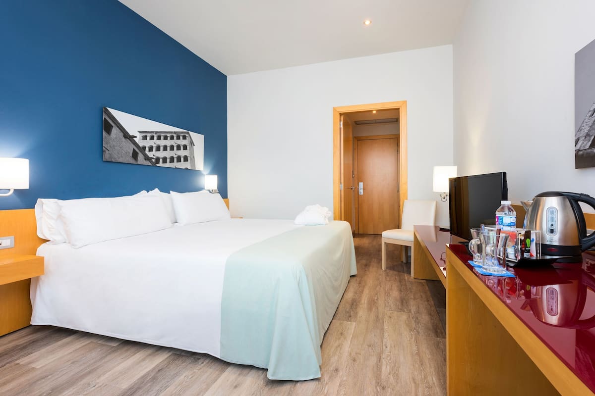A hotel room with a bed, a desk with a kettle, a chair, a TV, and a picture on the wall.