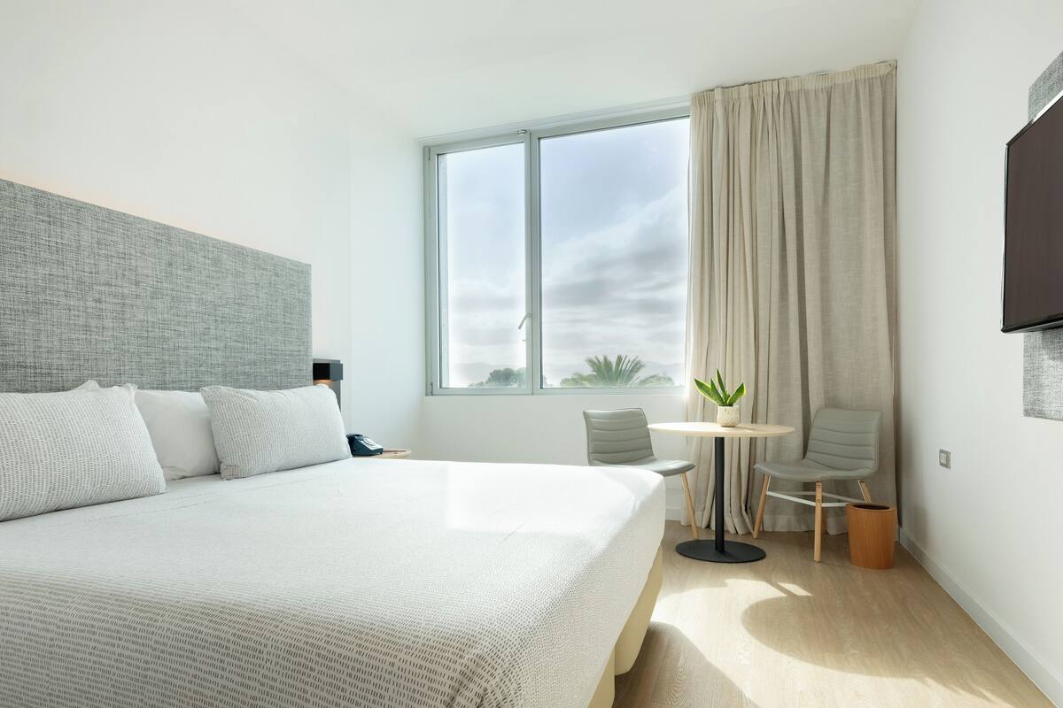 A modern hotel room with a large bed, a small round table with a plant, a grey armchair, and a TV mounted on the wall.