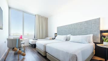 Habitación de hotel con dos camas, un escritorio, una silla, una mesita con una planta y un ventanal con cortinas.
