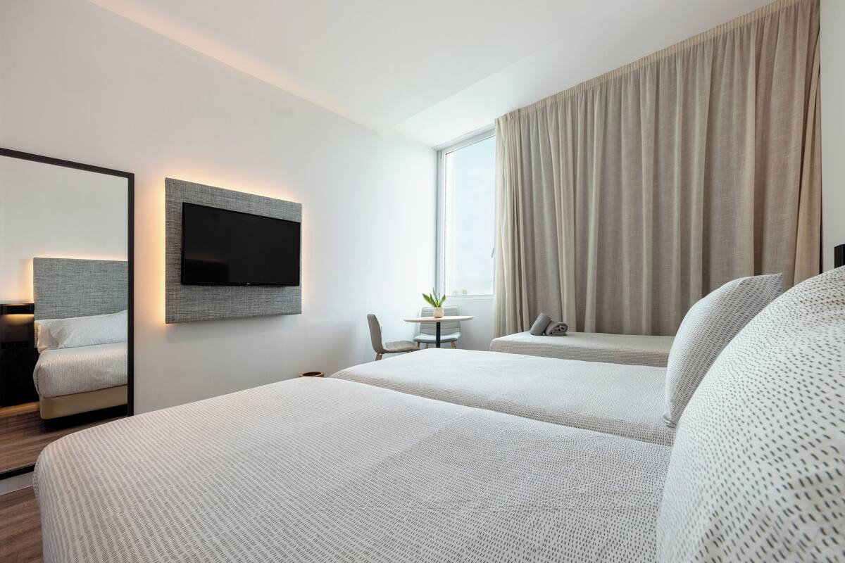 A hotel room with two beds, a TV mounted on the wall, a small table with a plant, and a large window with curtains.
