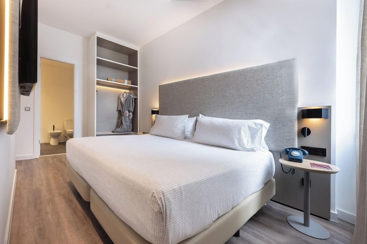 A modern hotel room with a large bed, built-in closet, bedside table with a phone, and a wall-mounted TV.