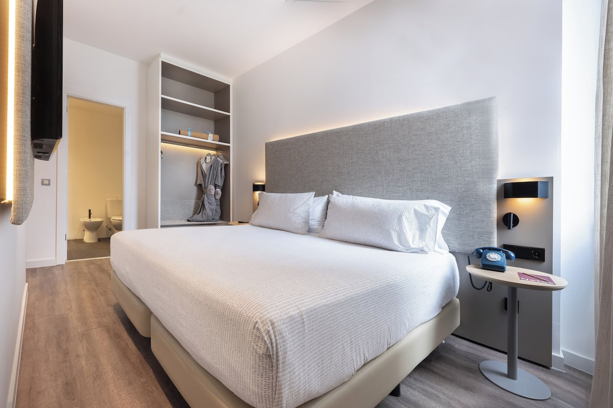 A modern hotel room with a large bed, a bedside table with a telephone, and a built-in wardrobe.