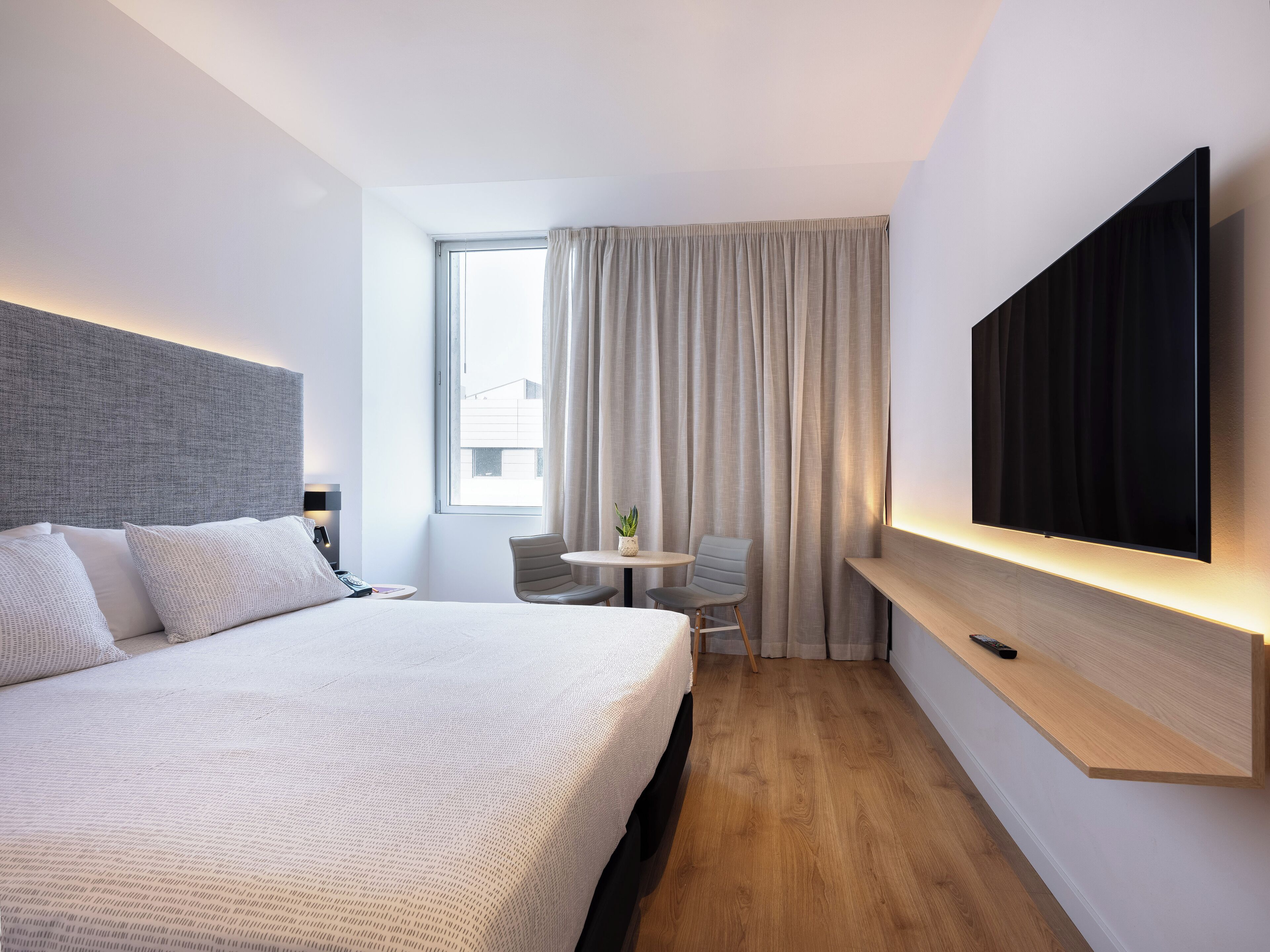 A modern hotel room with a large bed, a flat-screen TV mounted on the wall, a small table with a chair, and a window with curtains.