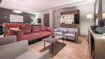 Una sala de estar moderna con un sofá rojo, sillones grises y una mesa de centro. Se observa un televisor, una estantería y una zona de cocina al fondo.