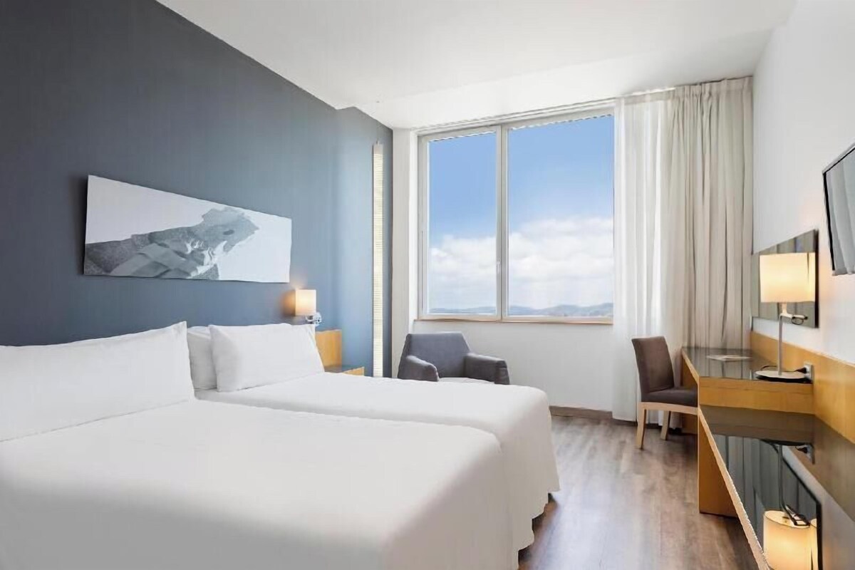 Una camera d'albergo con due letti, una scrivania, una sedia, una TV e un'ampia finestra con vista.