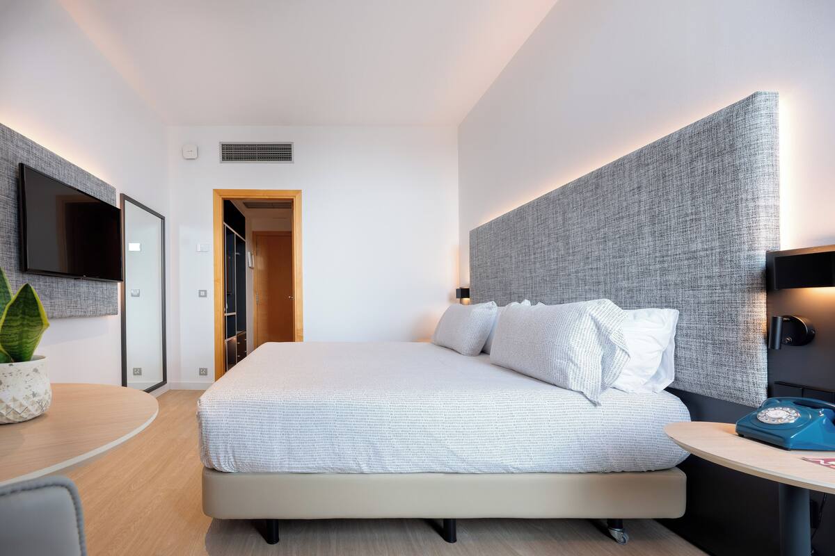 A modern hotel room with a large bed, a bedside table with a lamp, a TV mounted on the wall, and a plant on the side table.