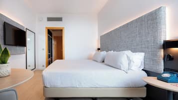 A modern hotel room with a large bed, a bedside table with a lamp, a TV mounted on the wall, and a plant on the side table.