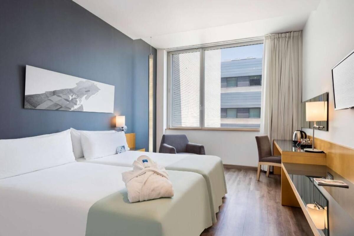 A hotel room with two beds, a desk, a chair, a window with blinds, and a painting on the wall.