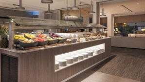 A buffet area with various fruits and bowls of food, a glass display case with plates, and modern pendant lighting. - INNSiDE by Meliá Barcelona Aeropuerto (El Prat de Llobregat)