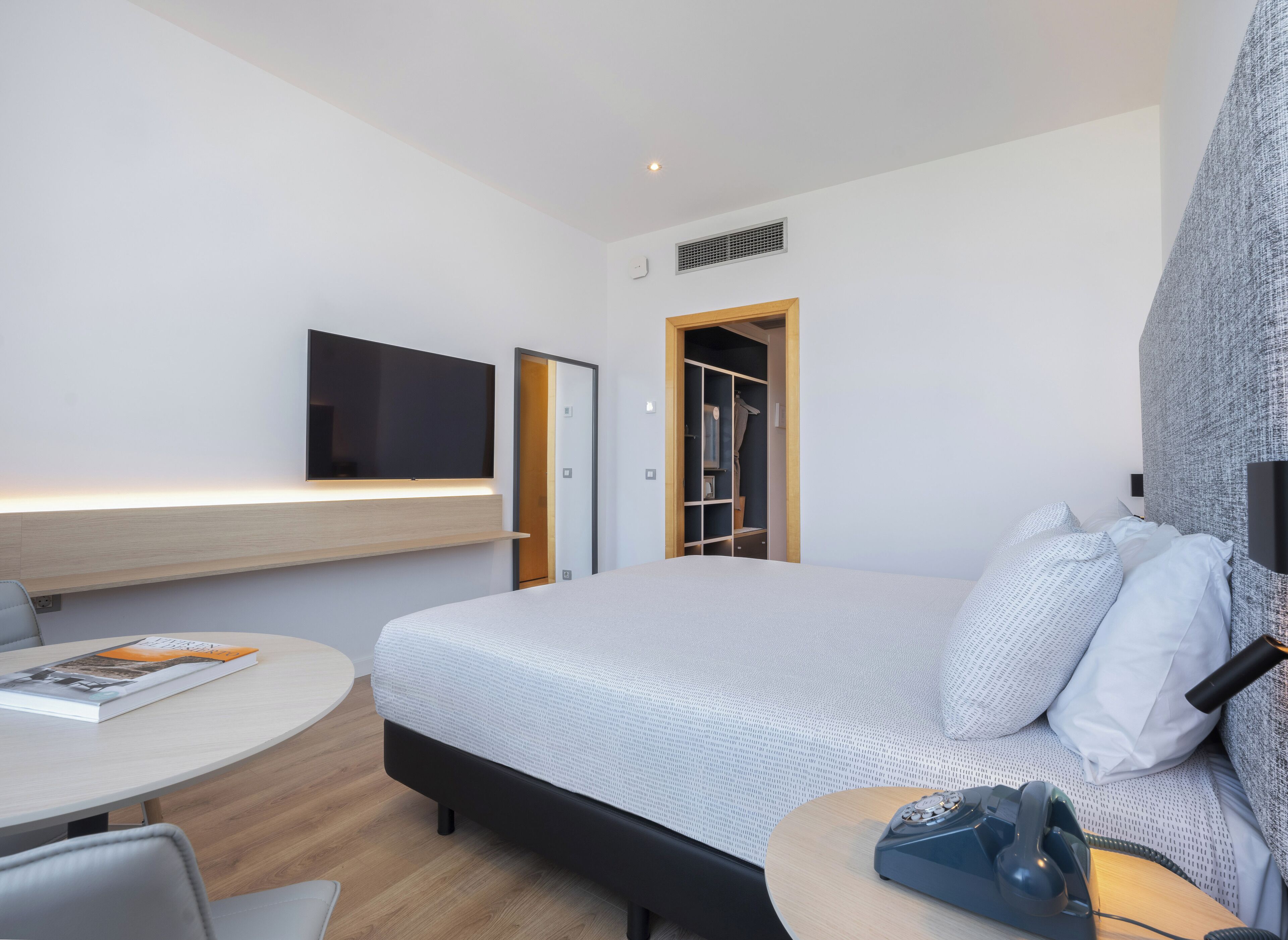 A modern hotel room with a bed, a desk, a chair, a television, and a wardrobe.
