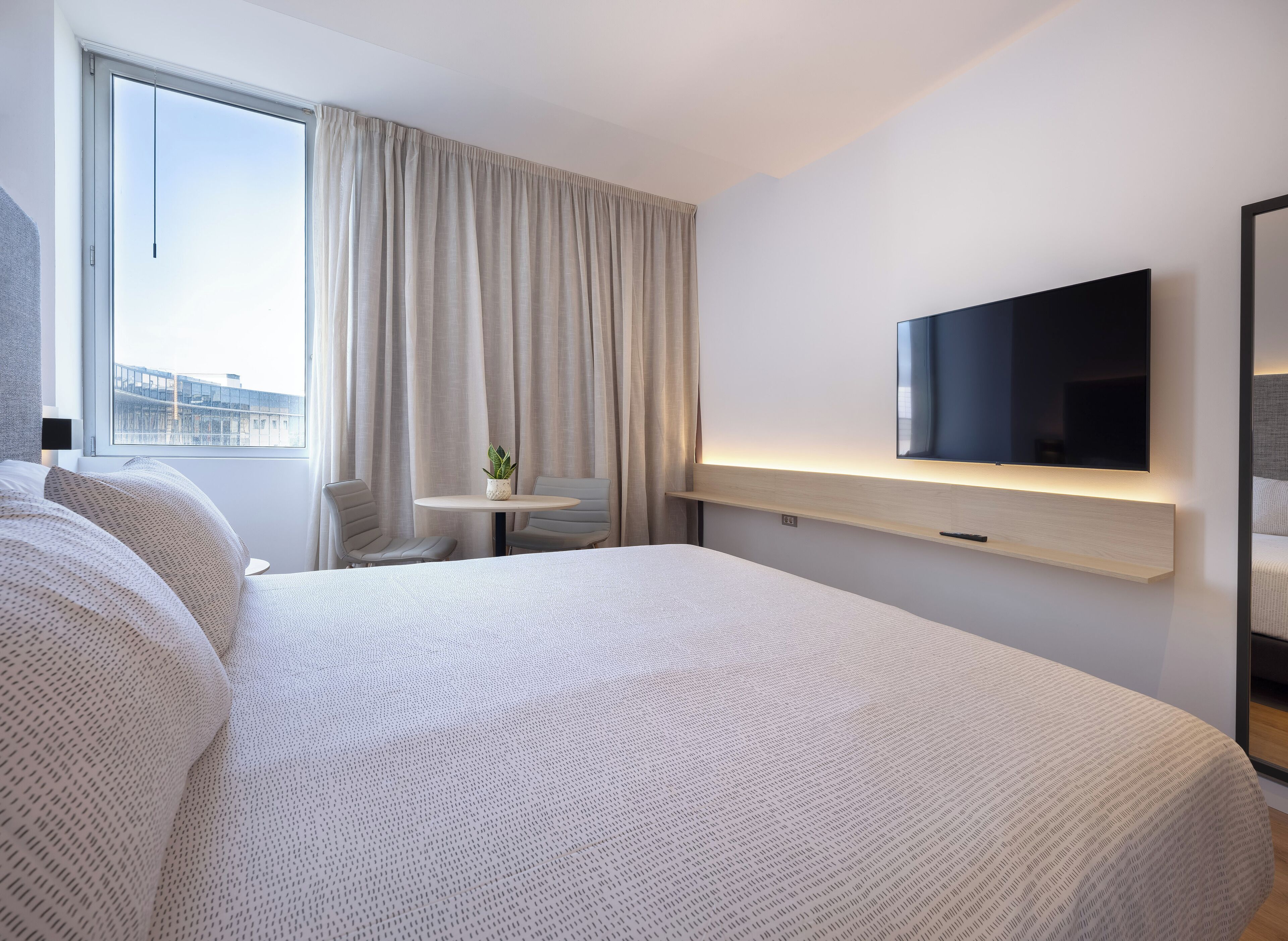 Una habitación de hotel moderna con una cama grande, un televisor de pantalla plana en la pared, una mesita redonda con una planta en maceta y vistas a la ciudad por la ventana.