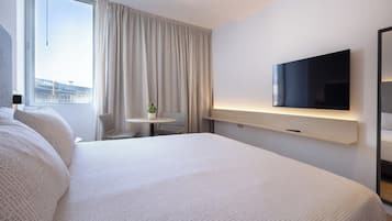 Una habitación de hotel moderna con una cama grande, un televisor de pantalla plana en la pared, una mesita redonda con una planta en maceta y vistas a la ciudad por la ventana.