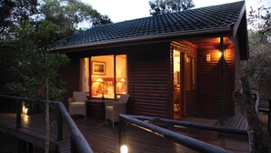A wooden cabin with a deck, a dining area with chairs and a table, and a lit interior.