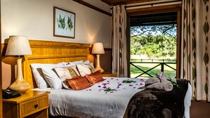 A hotel room with a wooden headboard, two bedside lamps, a bed with white bedding and a floral arrangement, and a view of greenery through the window.