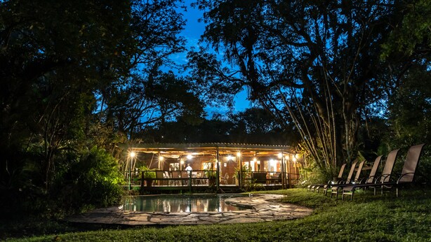 A wooden cabin with a pool, surrounded by trees and a well-lit patio area.