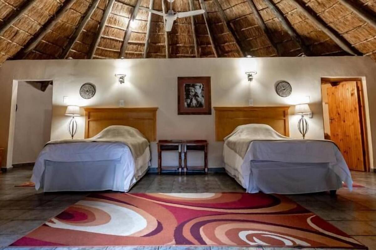 A room with a thatched roof, two beds, a small table, and a patterned rug.