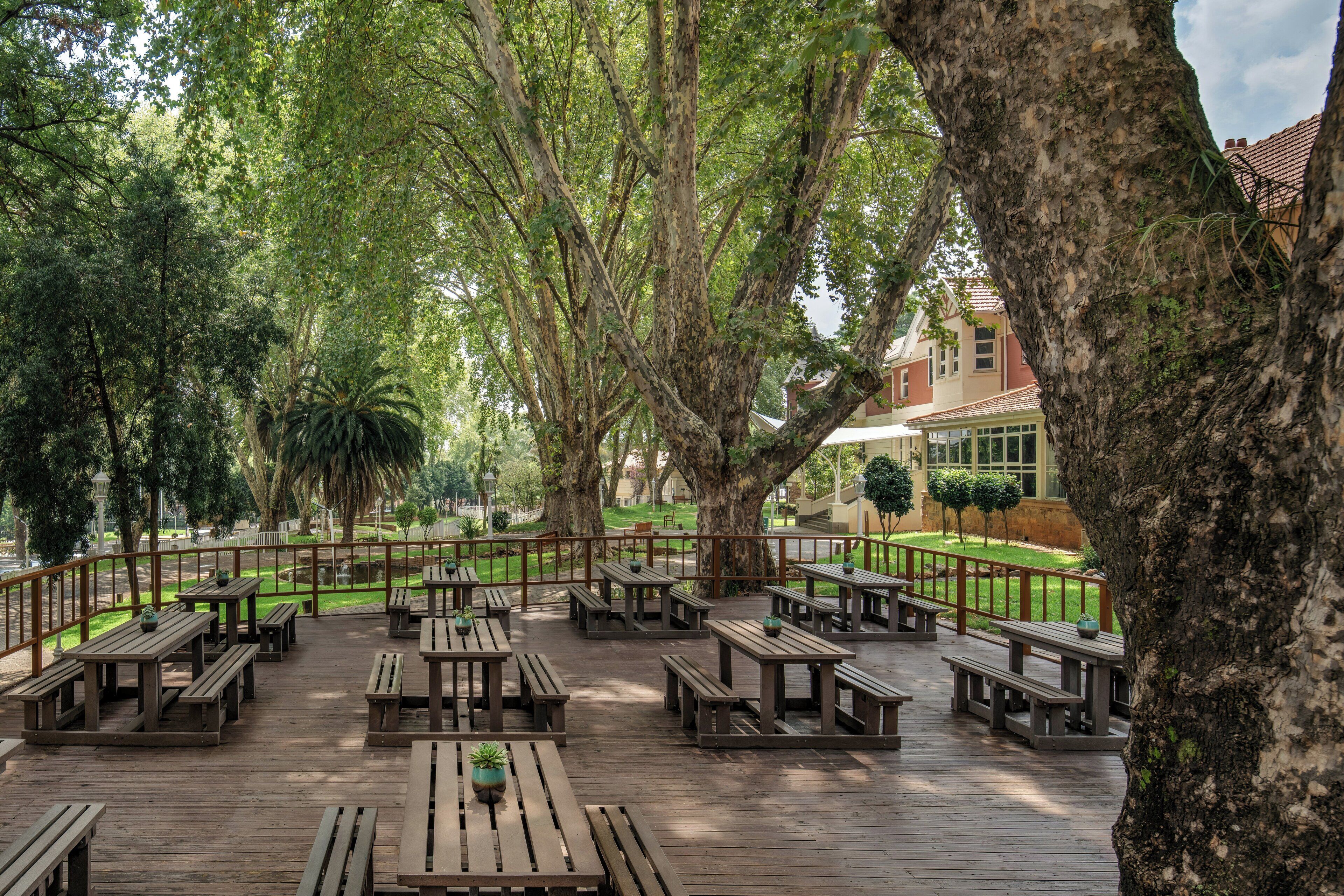 An outdoor seating area with wooden tables and benches, surrounded by trees and a view of a building.