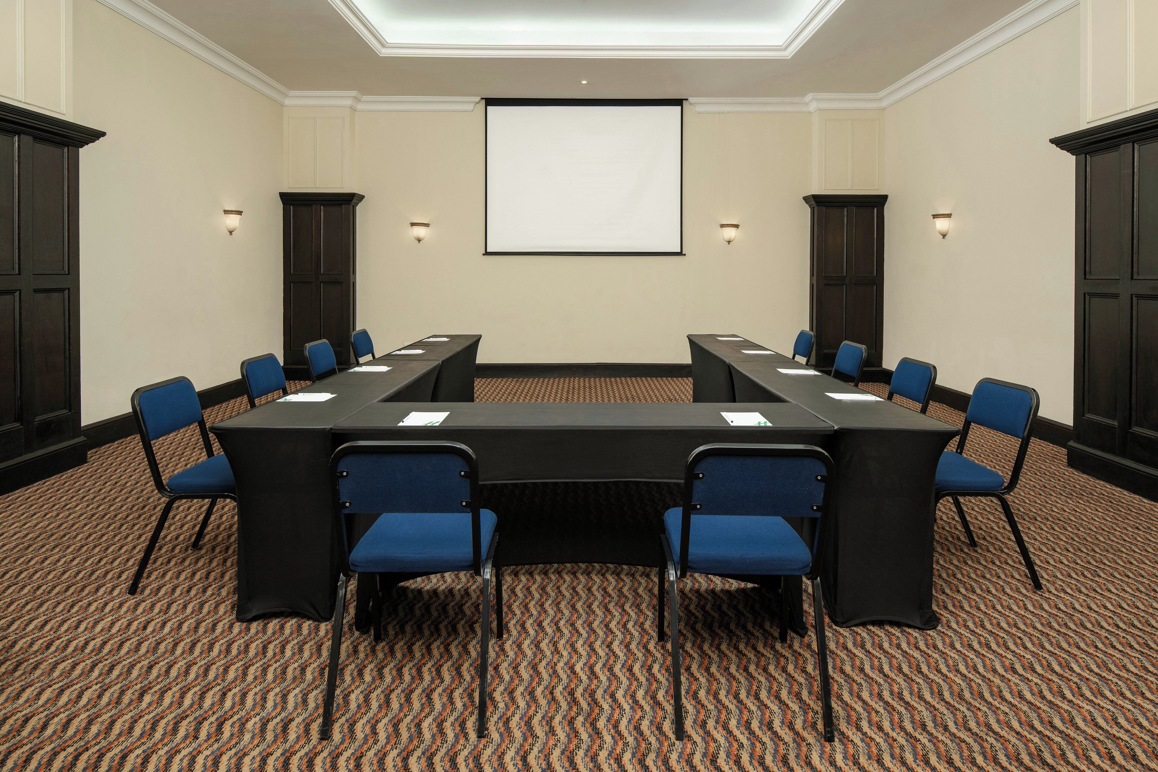 Un salón de conferencias con una gran mesa, sillas azules, una pantalla de proyector y armarios de madera oscura.