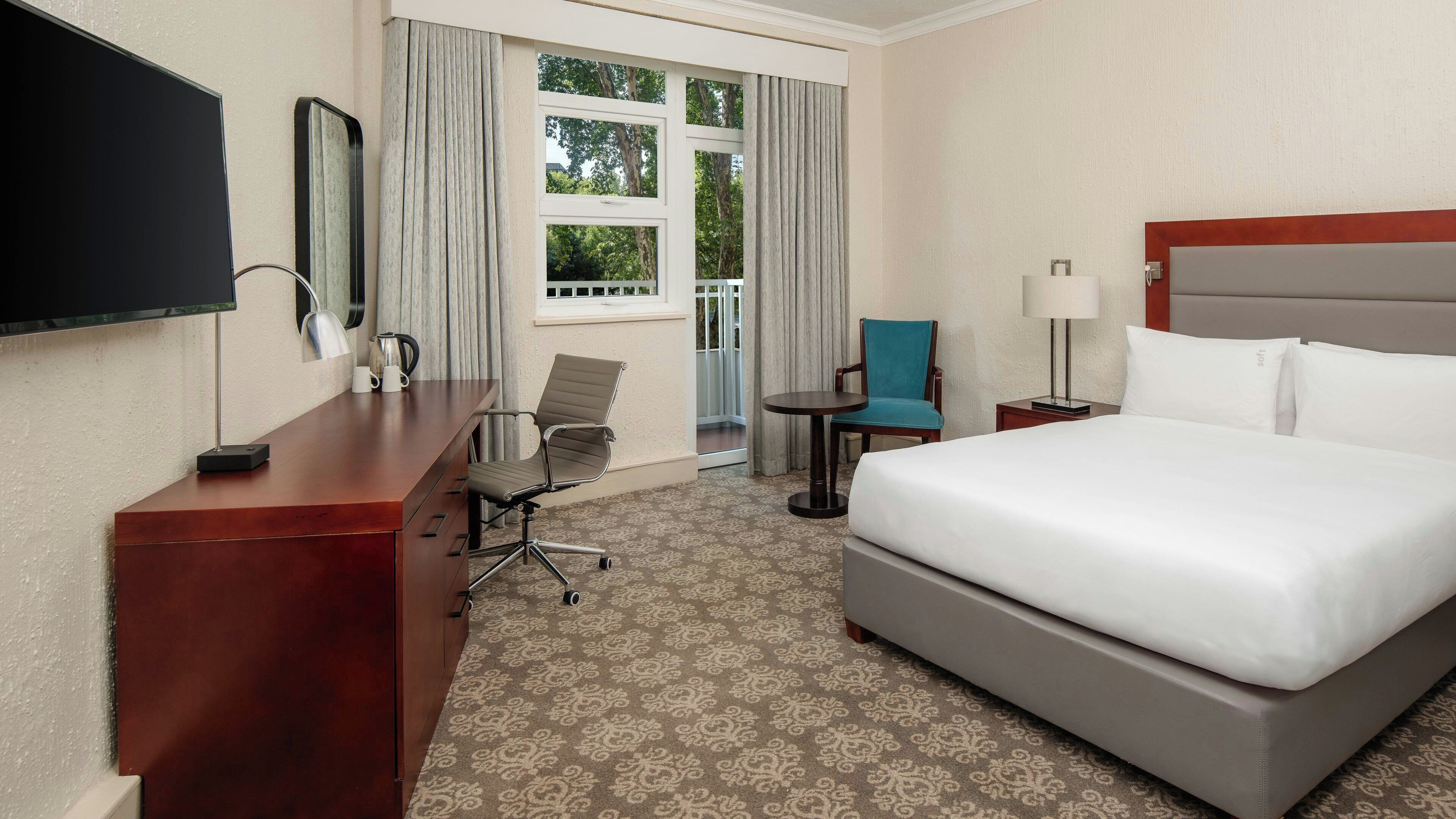 Una habitación de hotel con cama, escritorio, silla, televisión y ventana con cortinas.