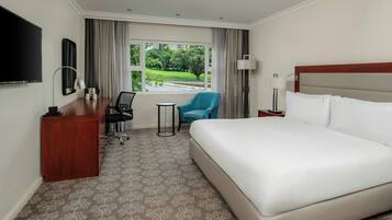 A hotel room with a large bed, a desk with a chair, a small table, a TV, and a window with a view of greenery.