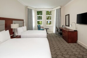 A hotel room with two beds, a desk, a chair, a TV, and a window with a view of trees.