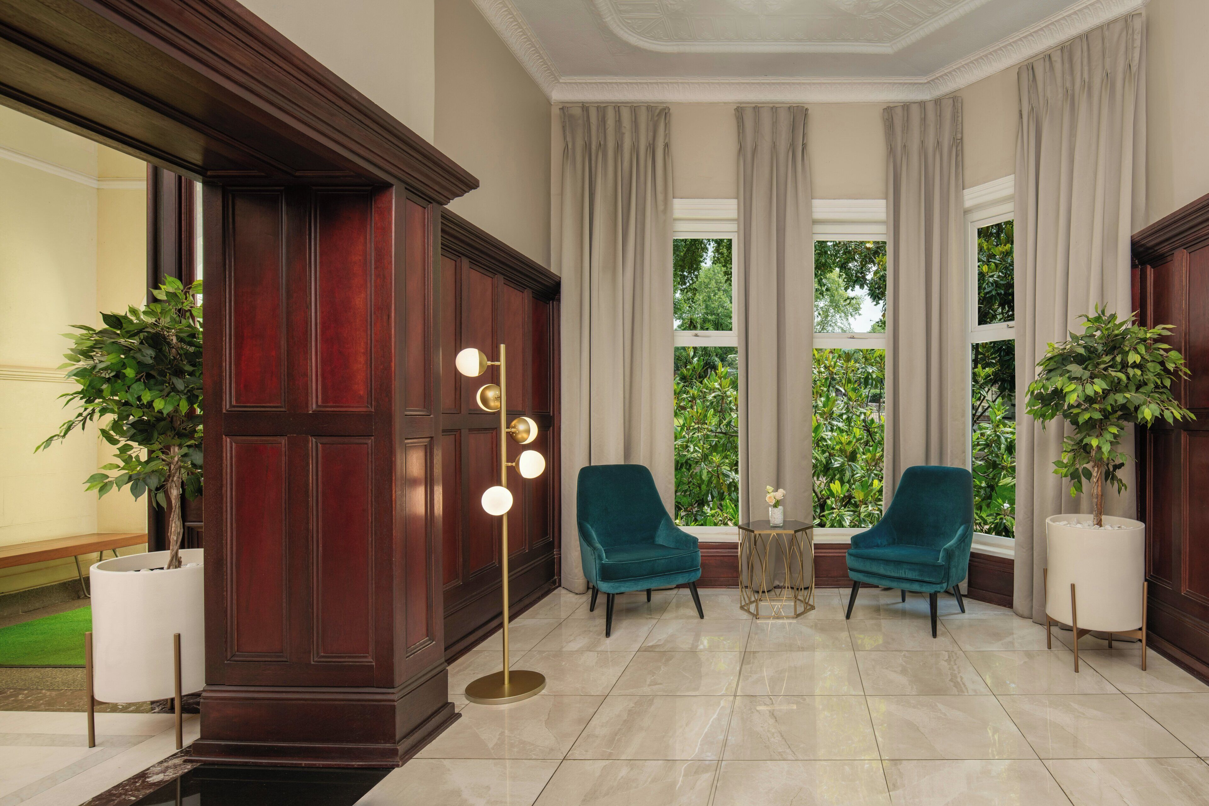 A room with a large wooden door, two chairs, a floor lamp, and a view of greenery through large windows.