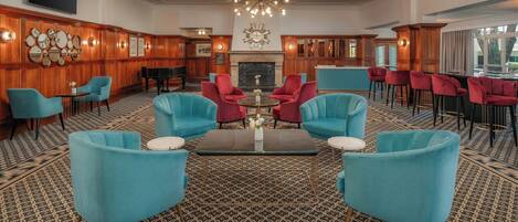 A lobby with a grand chandelier, a piano, and a fireplace. There are multiple seating areas with chairs and a coffee bar.