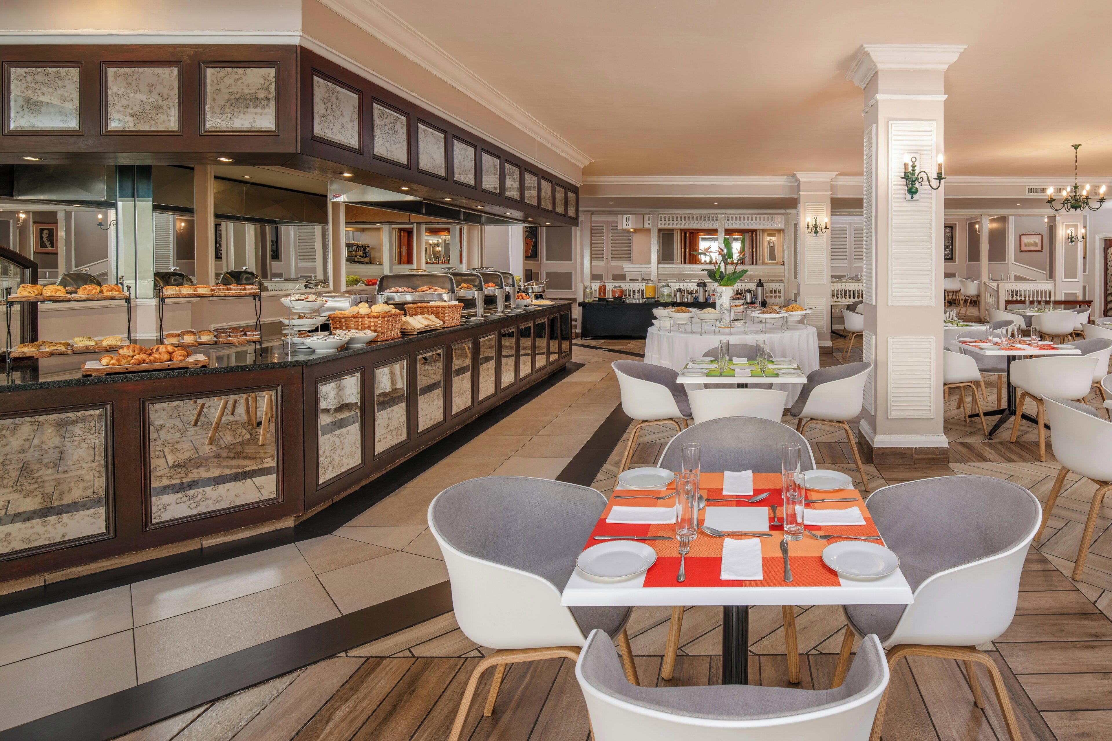 A hotel dining area with buffet-style breakfast options, white dining tables with orange placemats, and modern chairs.