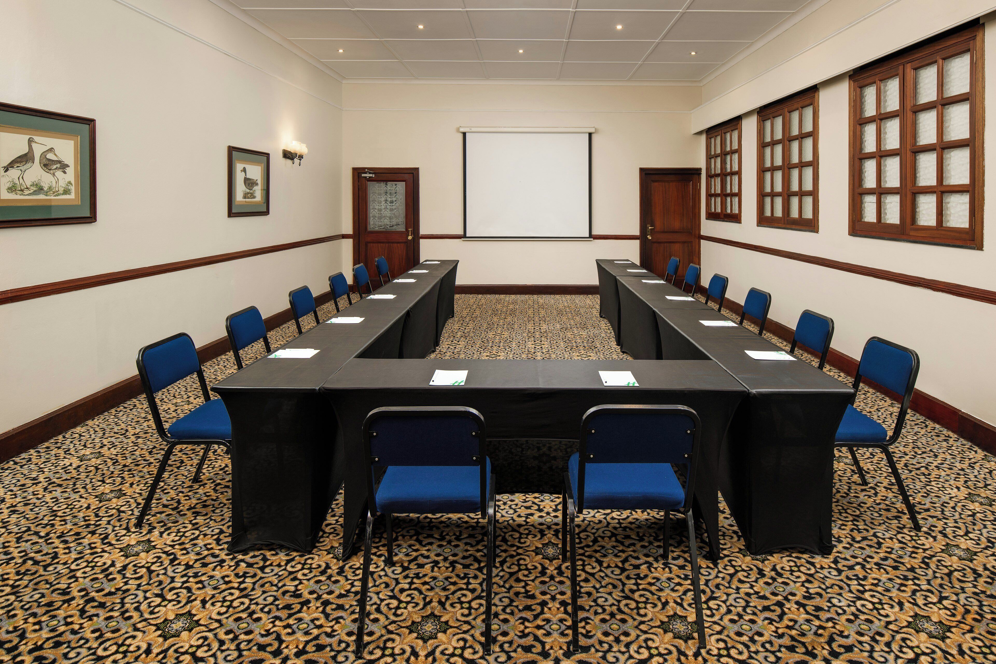 A conference room with a long table, blue chairs, a projector screen, and patterned carpet.