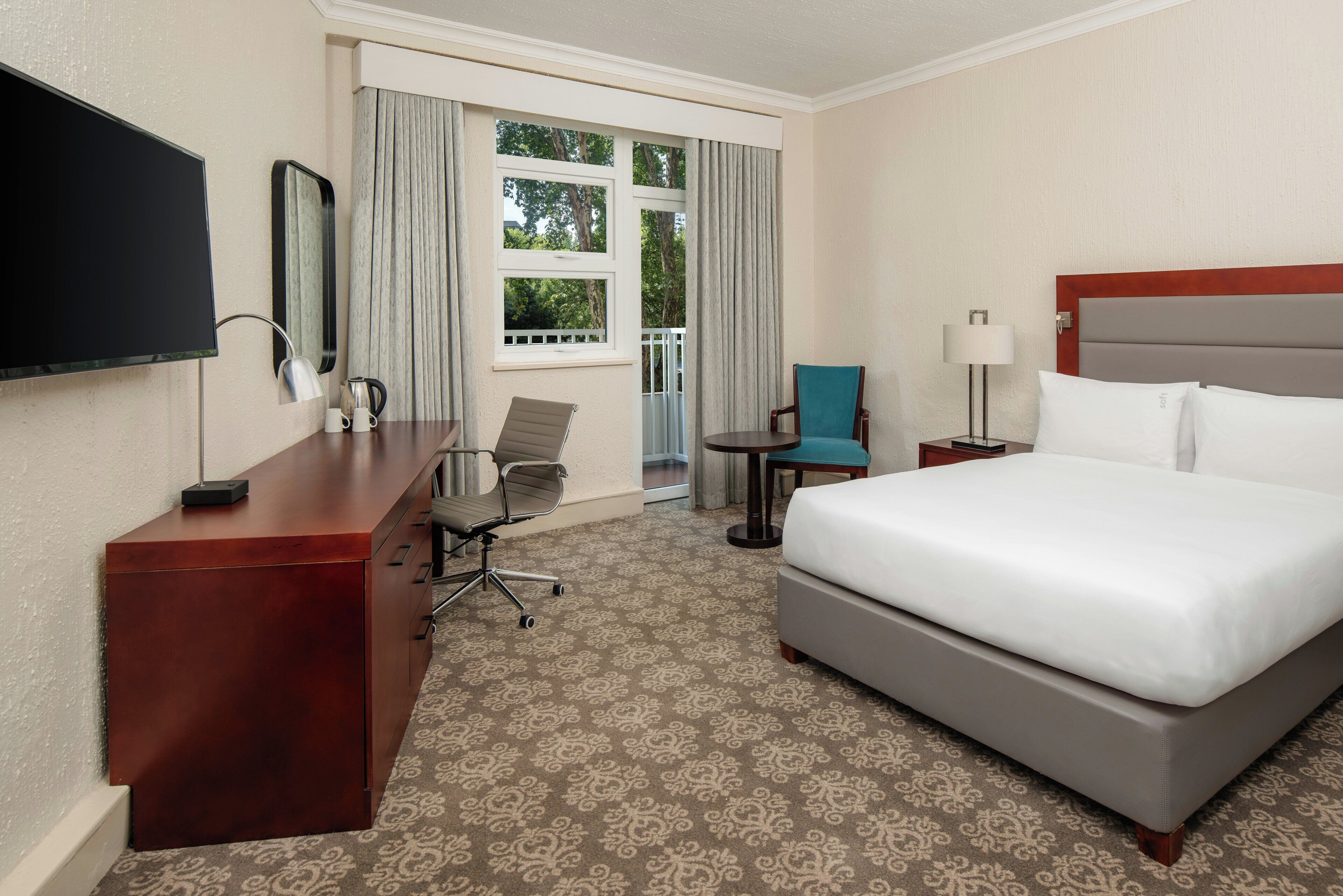 Quarto de hotel com cama, escrivaninha, cadeira, televisão e janela com cortinas.