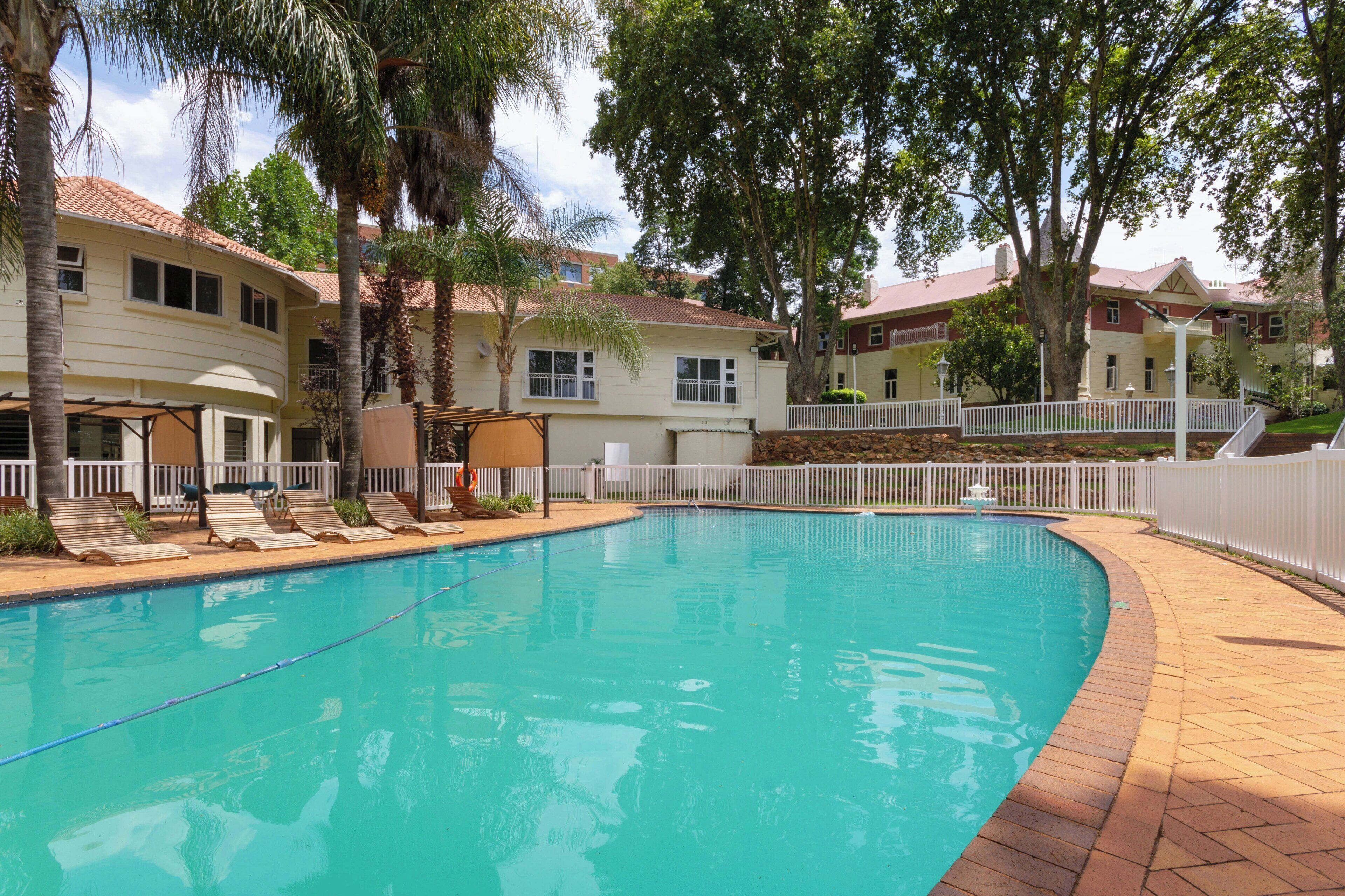 A swimming pool with lounge chairs, surrounded by a white fence and trees.