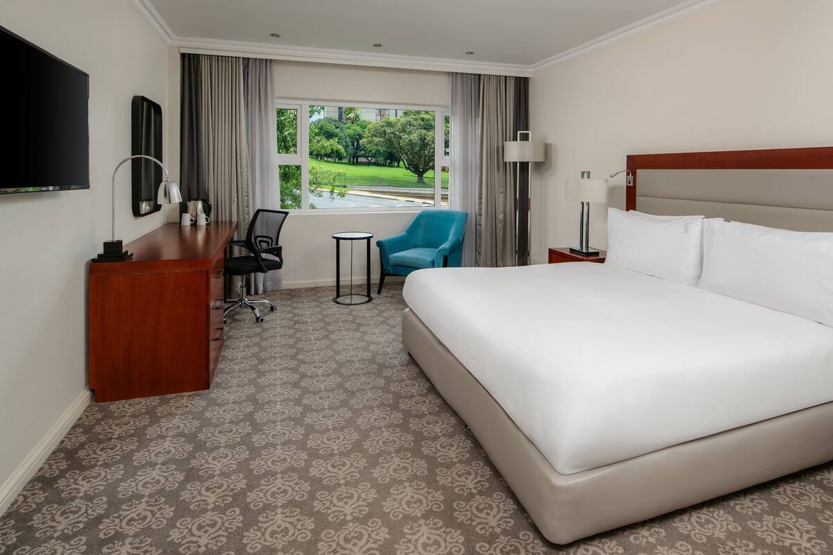 Une chambre d’hôtel avec un grand lit, un bureau avec une chaise, une petite table, une télévision et une fenêtre donnant sur un paysage verdoyant.