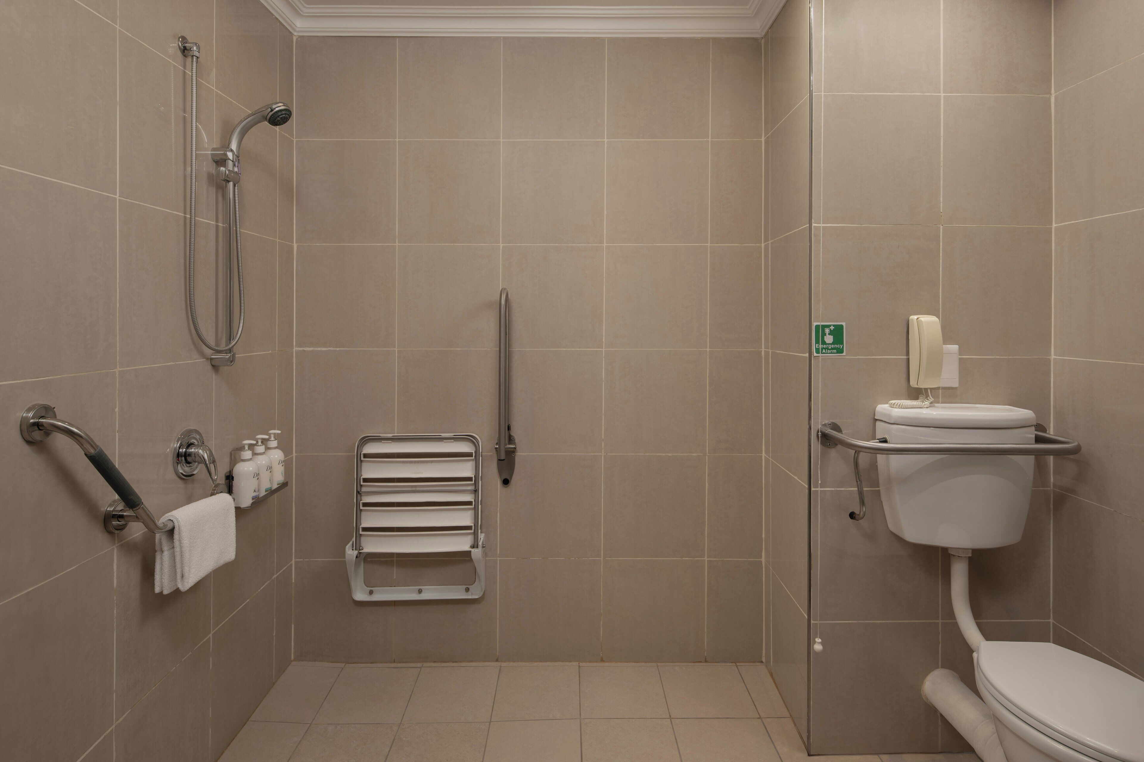 Ein Badezimmer mit Dusche, Toilette, Haltegriffen, einem Handtuchhalter und einem Notrufschild.