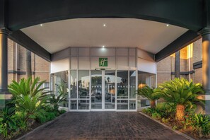 A hotel entrance with a glass facade, palm trees, and a brick building in the background. - Airport Grand Hotel and Conference Center (Boksburg)