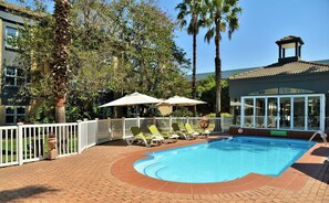 A swimming pool with lounge chairs and umbrellas, surrounded by a white fence and palm trees. - Airport Grand Hotel and Conference Center (Boksburg)