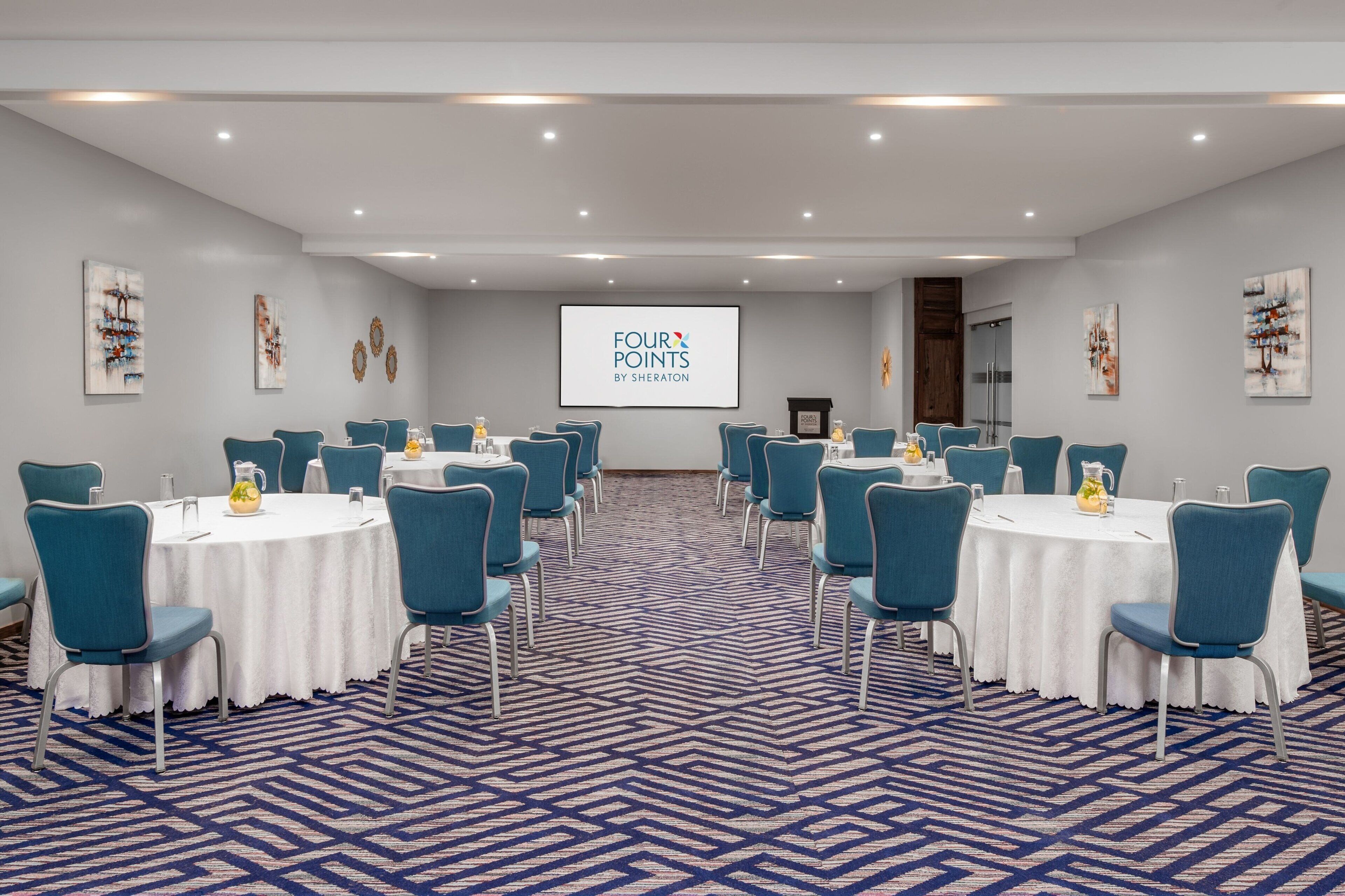 Una sala conferenze con tavoli rotondi, sedie color acquamarina e un grande schermo che mostra "FOUR POINTS BY SHERATON".
