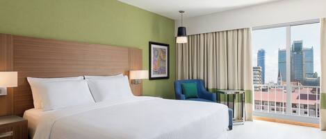A hotel room with a large bed, a desk, a chair, a window with a city view, and a framed artwork on the wall.