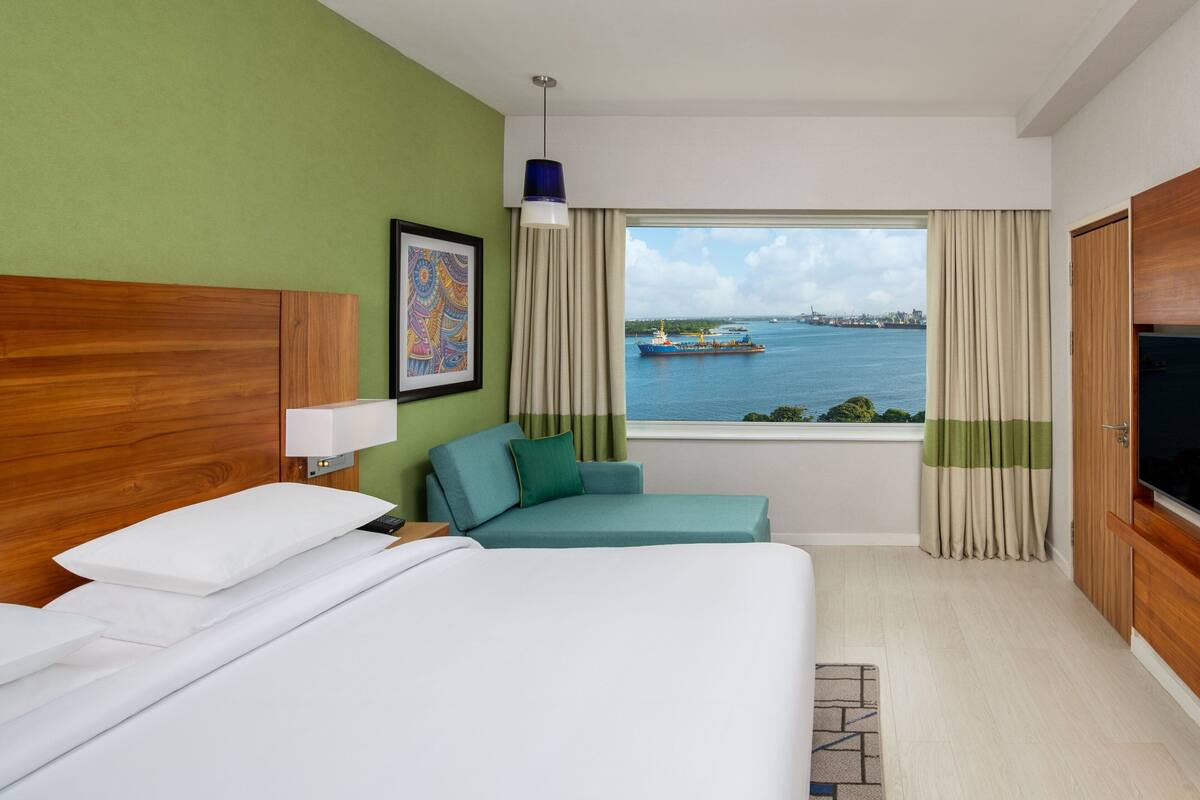 Quarto de hotel com uma cama, um sofá, uma televisão e uma janela com vista para a água e um navio.