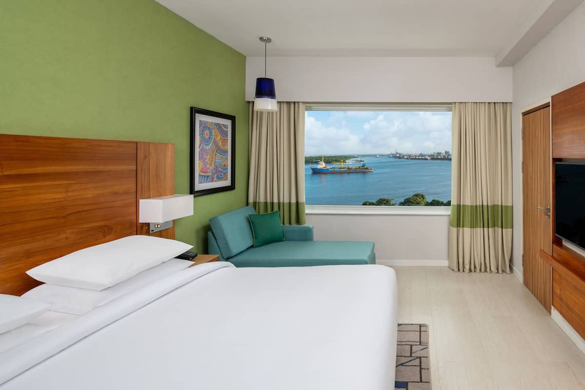 A hotel room with a bed, a sofa, a TV, and a window with a view of a body of water and a ship.
