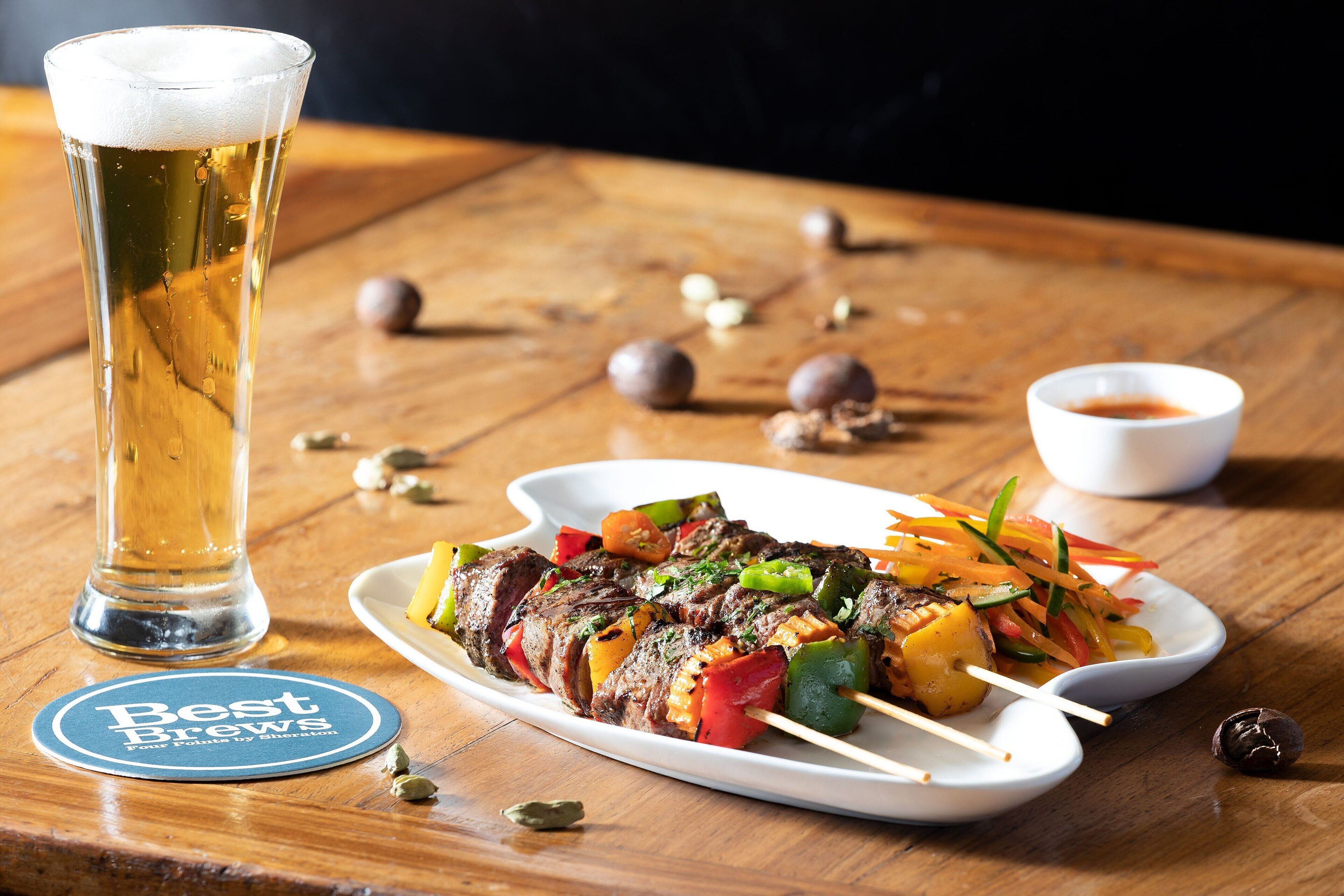 A plate of skewered meat and vegetables, a glass of beer, and a coaster with text on a wooden table.