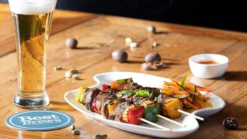A plate of skewered meat and vegetables, a glass of beer, and a coaster with text on a wooden table.