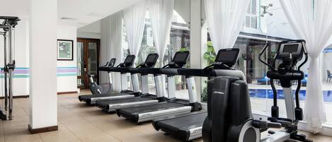 A gym with multiple treadmills and a stationary bike, large windows with curtains, and a view of a pool.