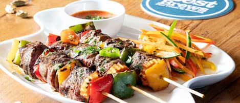 Un plato con brochetas de carne y verduras, un vaso alto de cerveza y un posavasos con la inscripción 'Best Brews'.