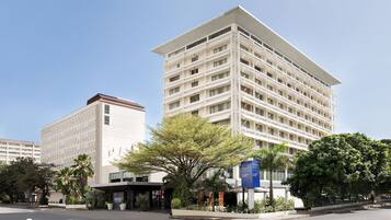 Um hotel de vários andares com fachada moderna, área de entrada arborizada e céu claro.