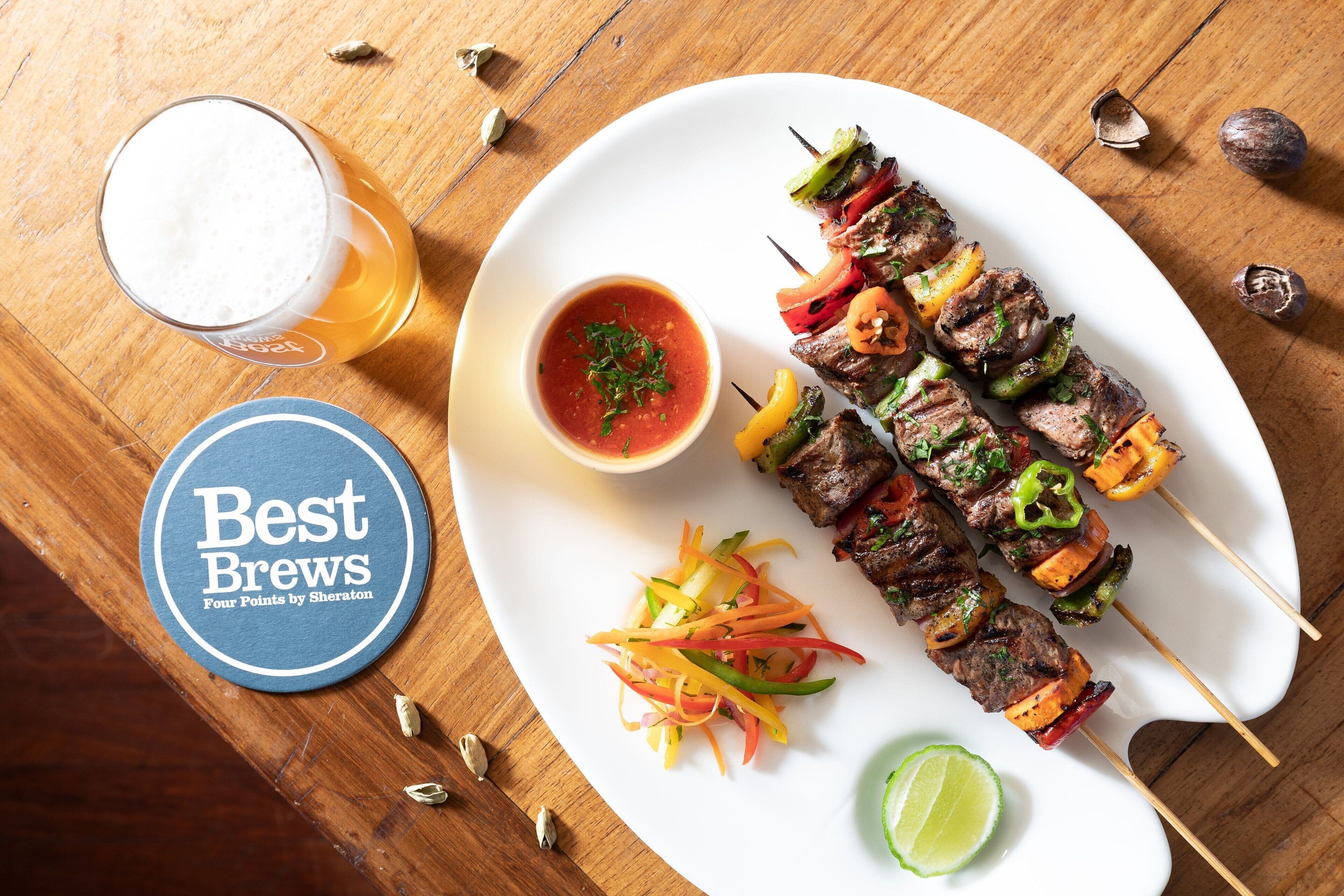 A plate of skewered meat with vegetables, a glass of beer, and a small bowl of sauce on a wooden table.