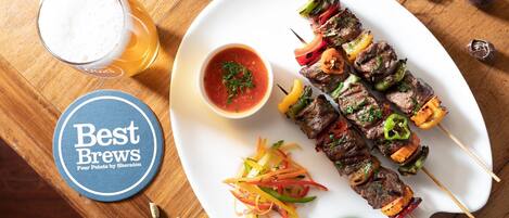 A plate of skewered meat with vegetables, a glass of beer, and a small bowl of sauce on a wooden table.